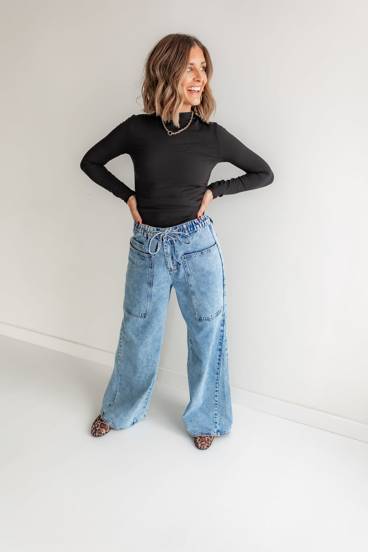 Woman wearing a black long-sleeve top and wideleg blue jeans against a white background