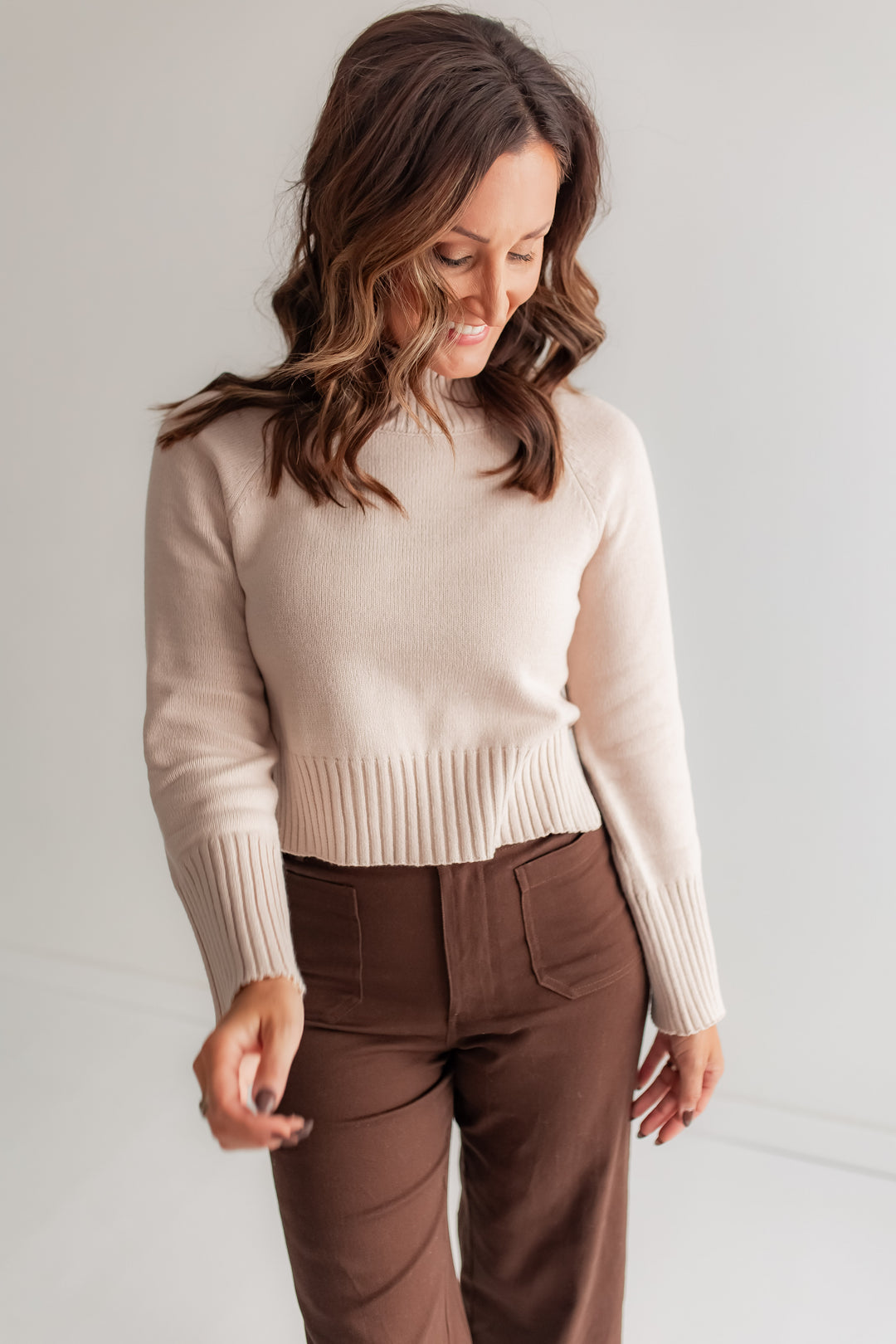 A woman poses in a creamy white, semi-cropped mockneck sweater with ribbed cuffs and hem. Brown Pants