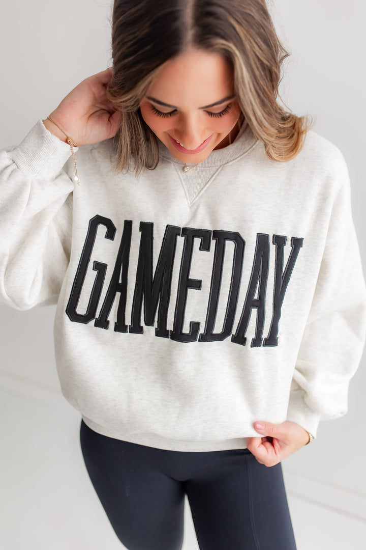 A close-up shot of a heather grey crewneck sweatshirt with "GAMEDAY" embroidered in black on the front.