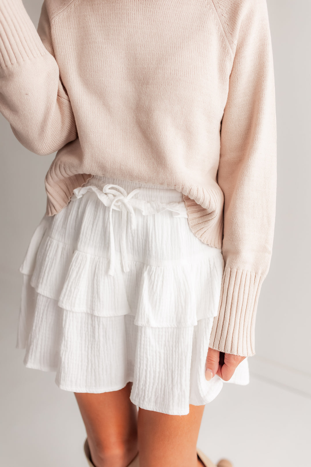  A woman stands outdoors, wearing a white tiered ruffle mini skirt with a tied waist, paired with a cozy knit sweater and tall boots.