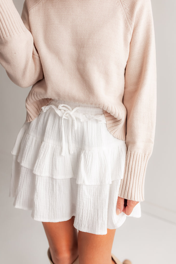  A woman stands outdoors, wearing a white tiered ruffle mini skirt with a tied waist, paired with a cozy knit sweater and tall boots.