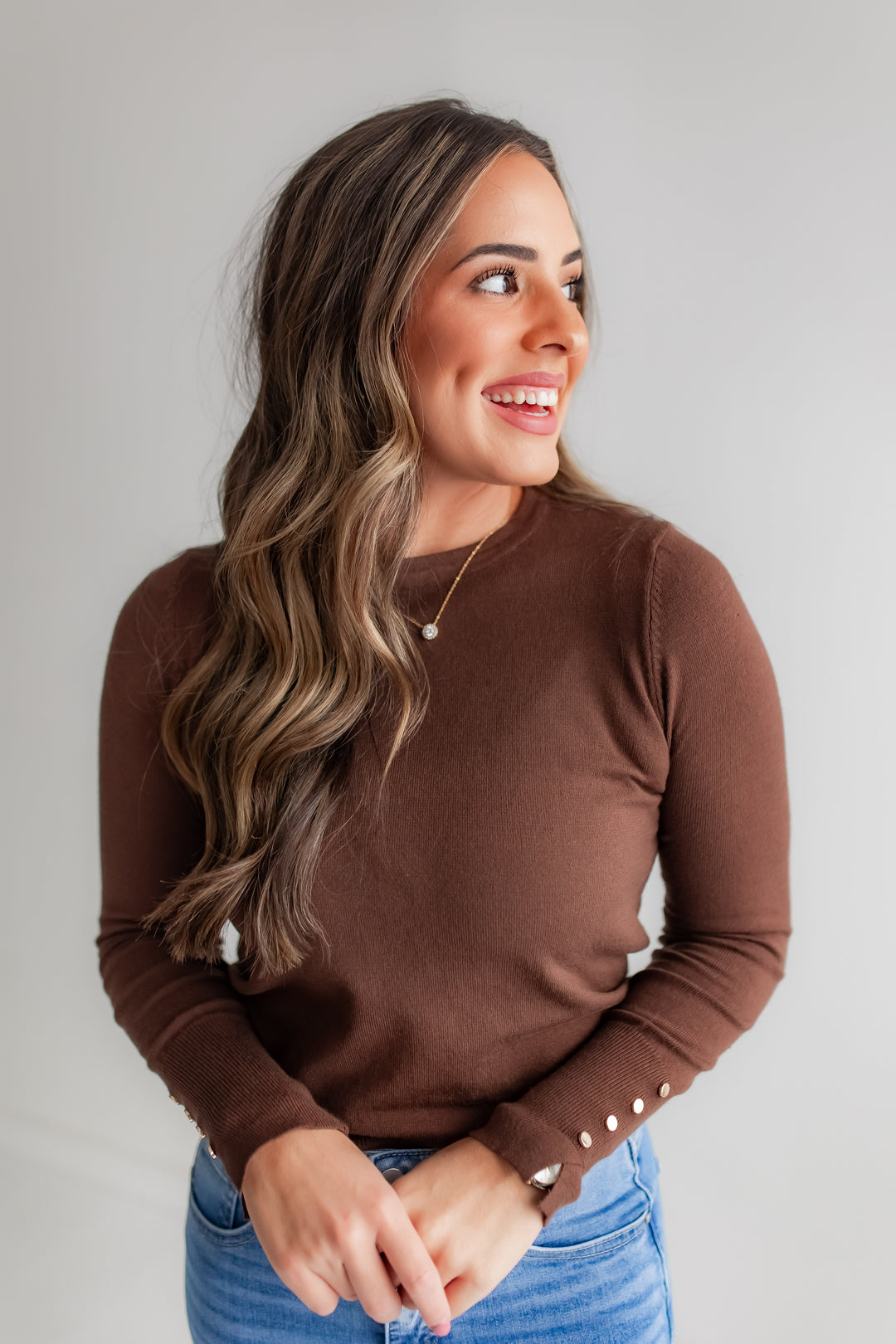 A woman wears a rich brown long-sleeve top with gold button details on the sleeves.