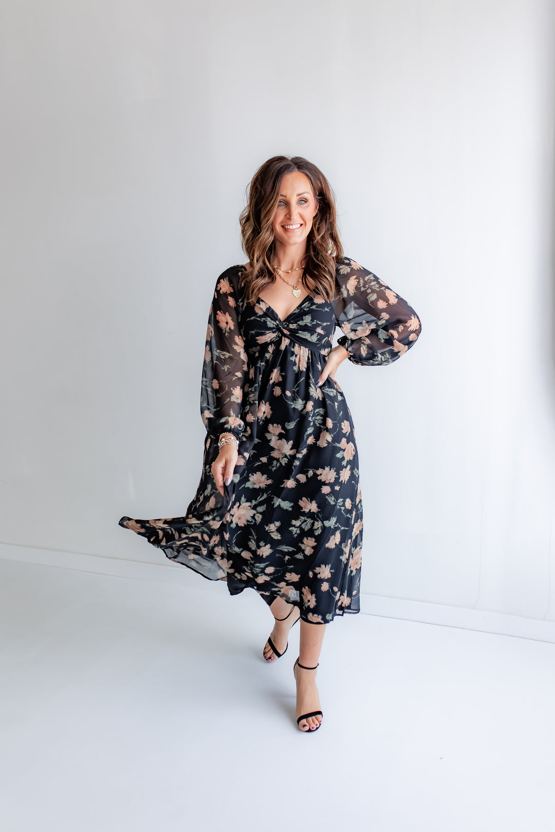  A woman stands outdoors, wearing a black maxi dress with a floral print, sheer long sleeves, and a twisted bodice.
