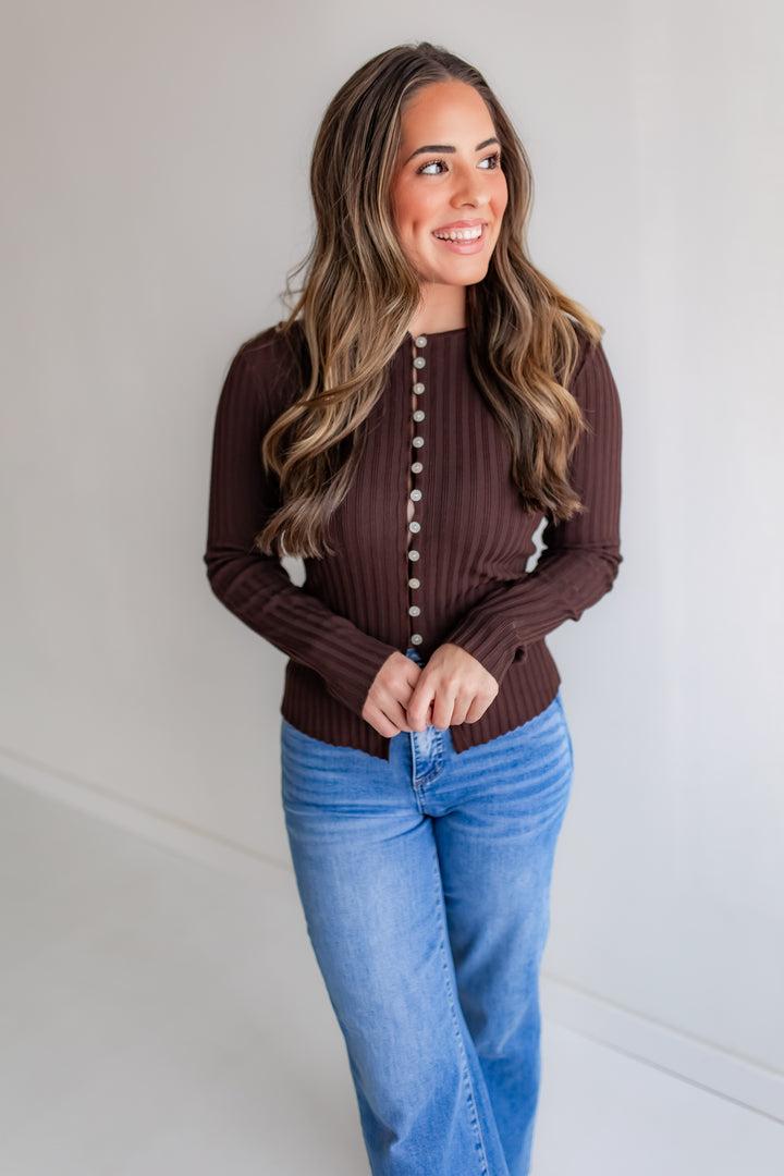 A woman wears a brown long-sleeve crewneck top with full-length button details and a ribbed texture.