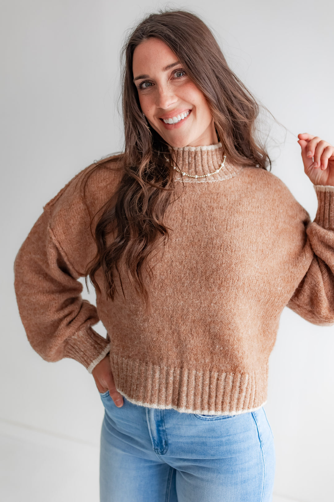 A woman poses in a cozy, oversized tan mockneck sweater with cream-colored piping on the shoulders.