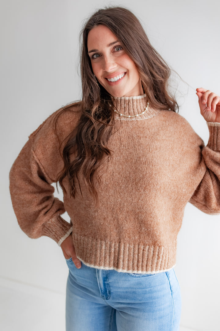 A woman poses in a cozy, oversized tan mockneck sweater with cream-colored piping on the shoulders.