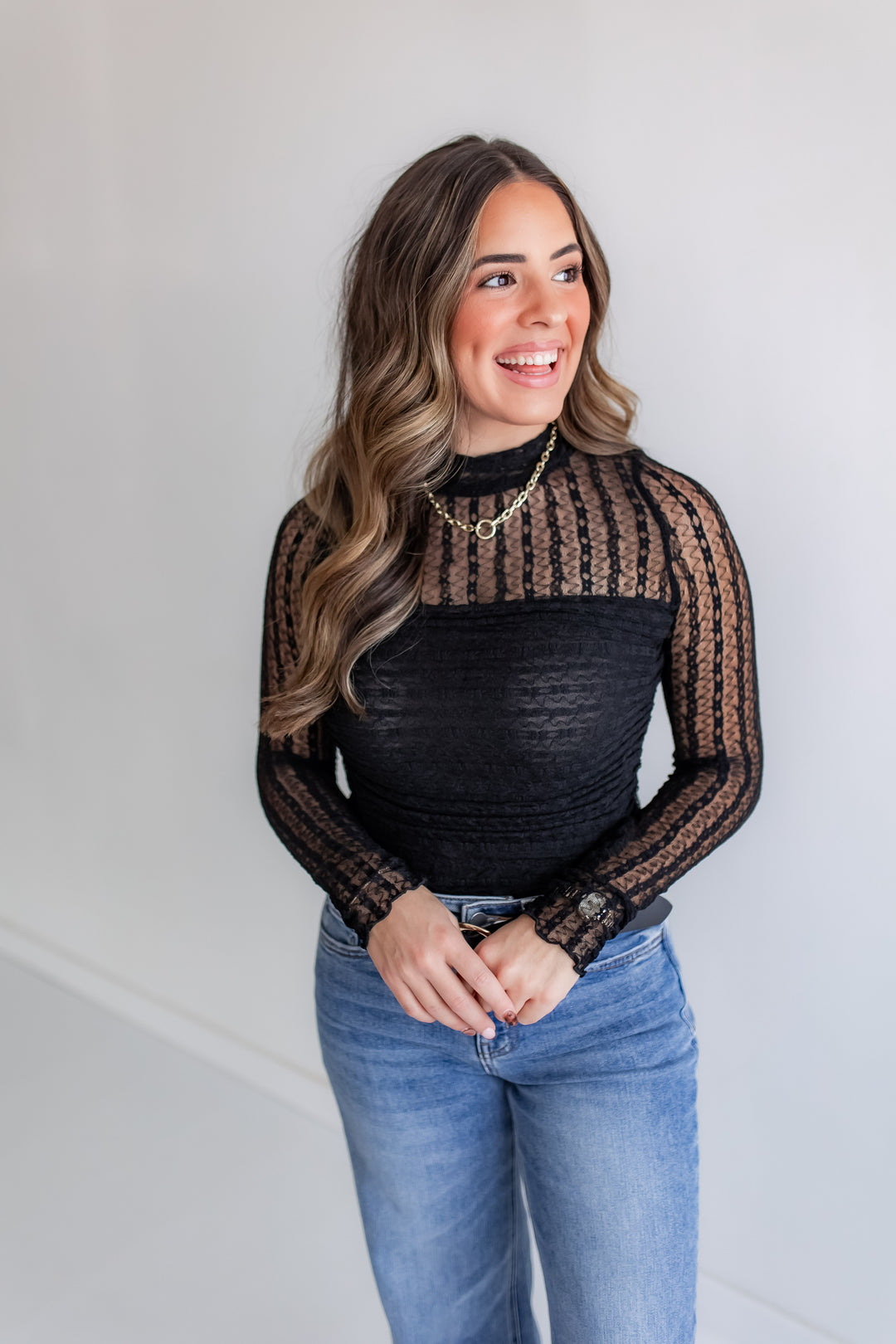 A woman poses in a black lace mockneck bodysuit with ruched detailing on the torso and sheer sleeves.