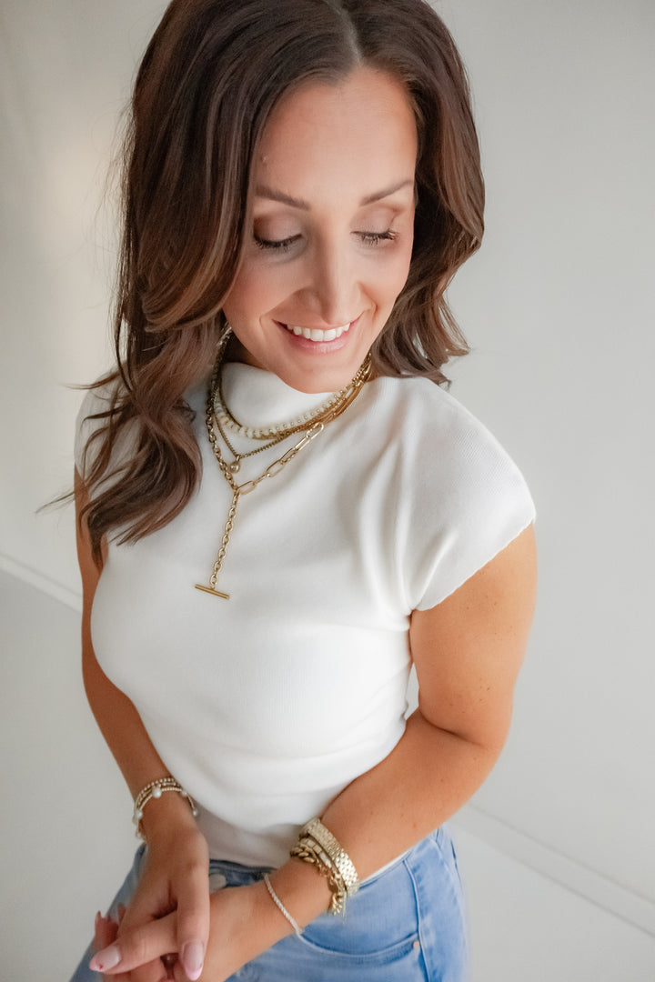 Woman wearing a white top and blue jeans with layered necklaces and bracelets on a plain background