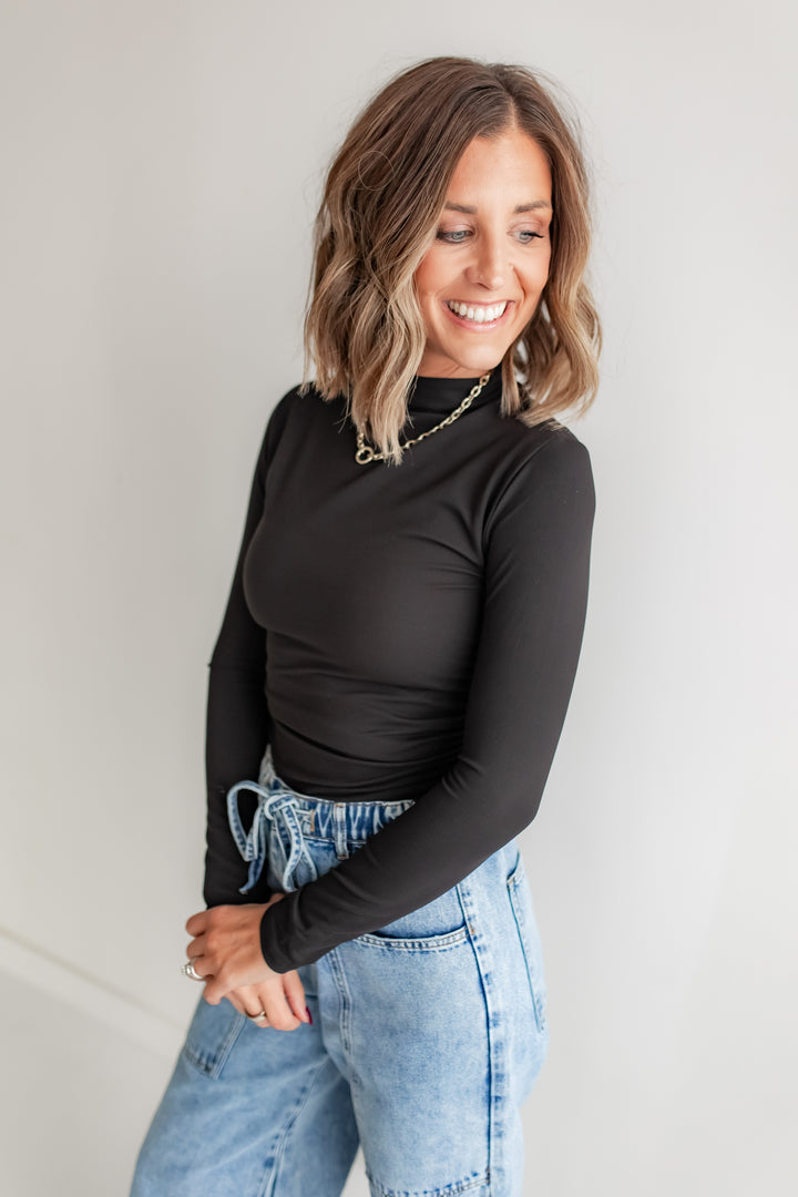 Woman wearing a black long-sleeve top and blue jeans against a plain background