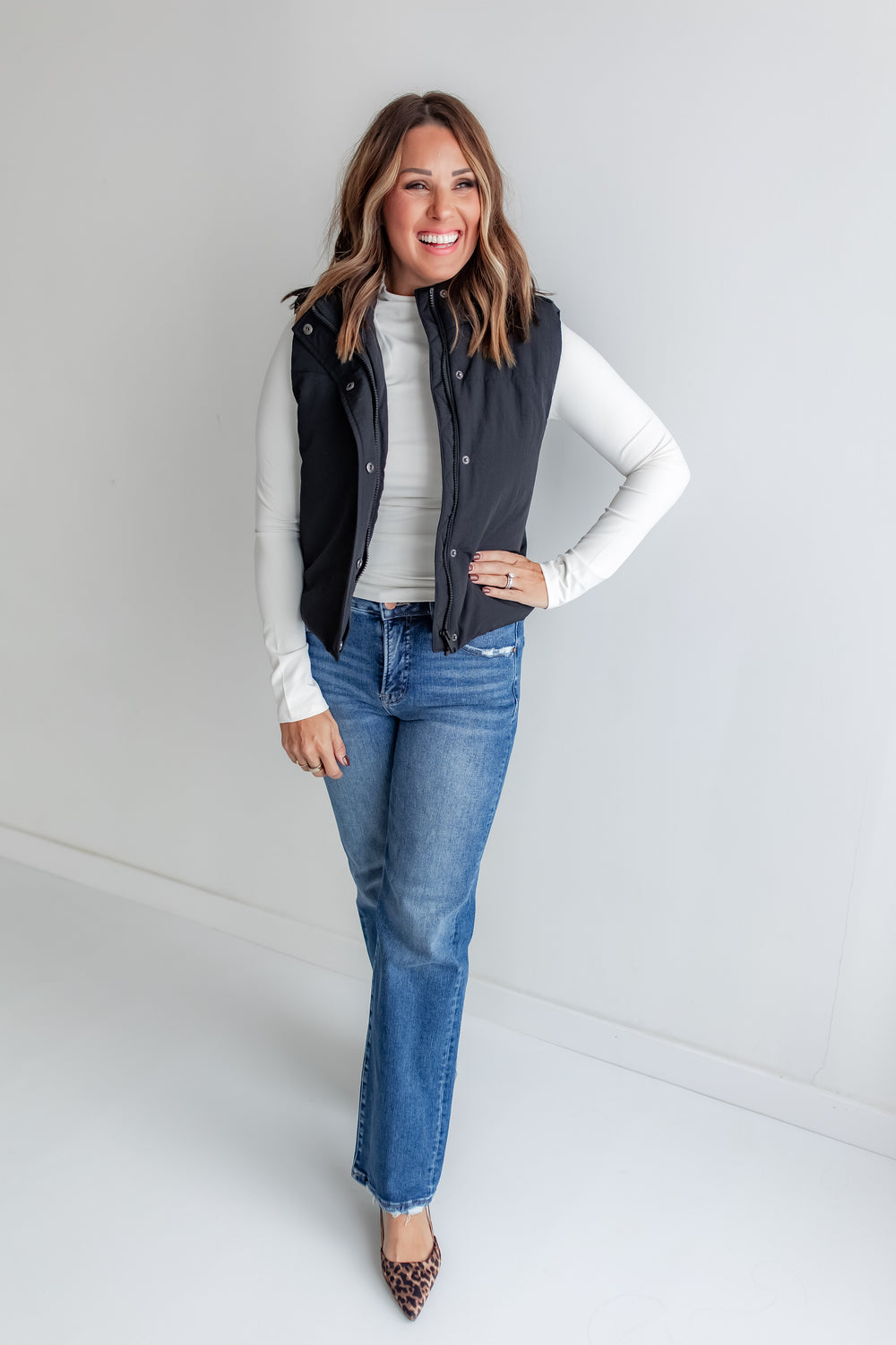 Woman wearing a black puffer vest with buttons, white long-sleeve shirt, blue jeans, and leopard print shoes