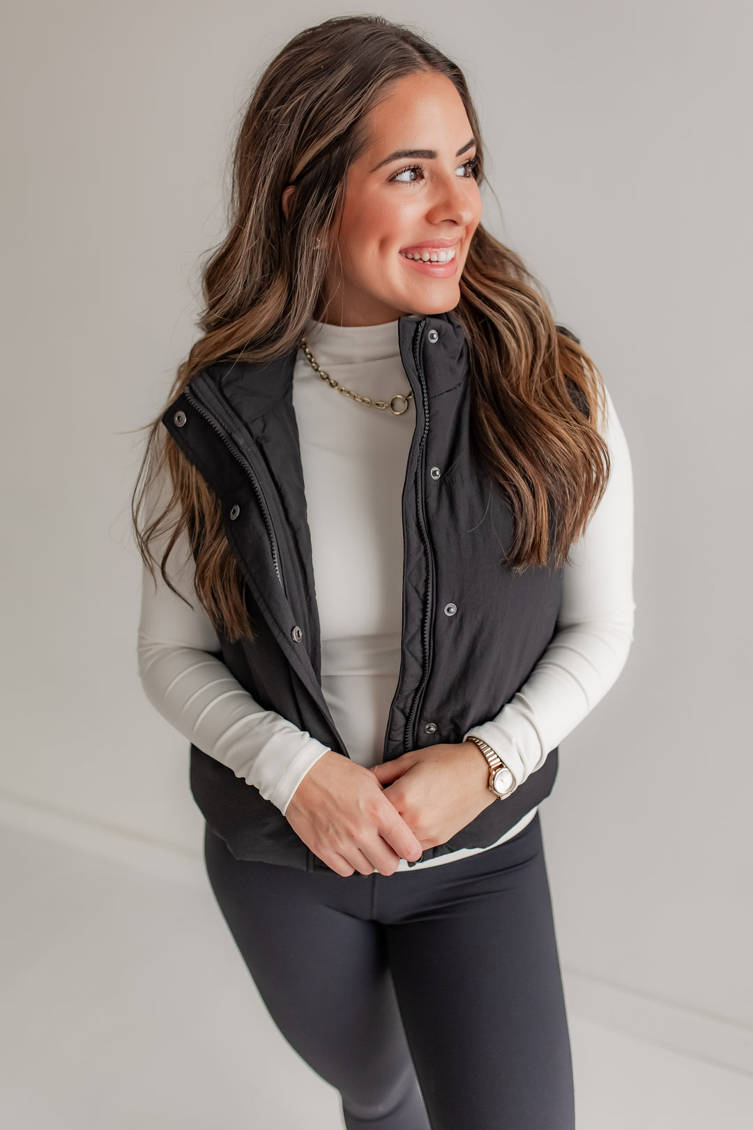 Woman wearing a black puffer vest over a white long-sleeve shirt and leggings on a plain background