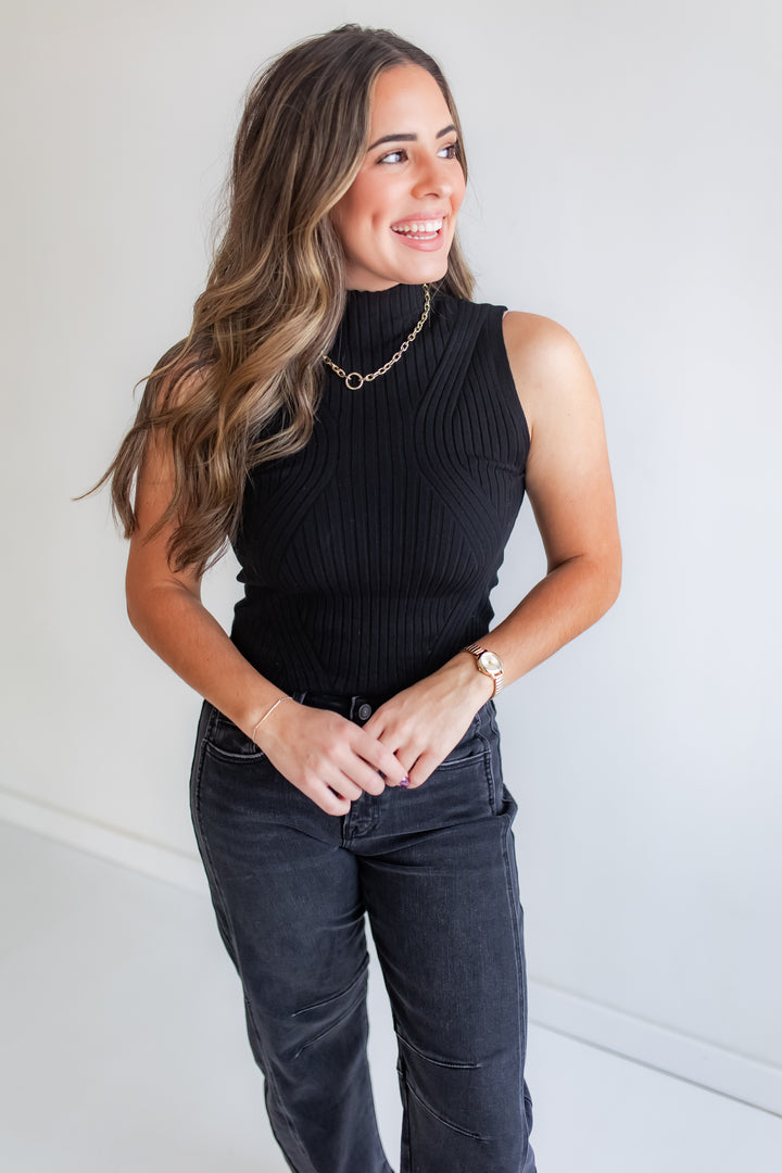 Woman wearing a black sleeveless mock neck top and dark jeans.