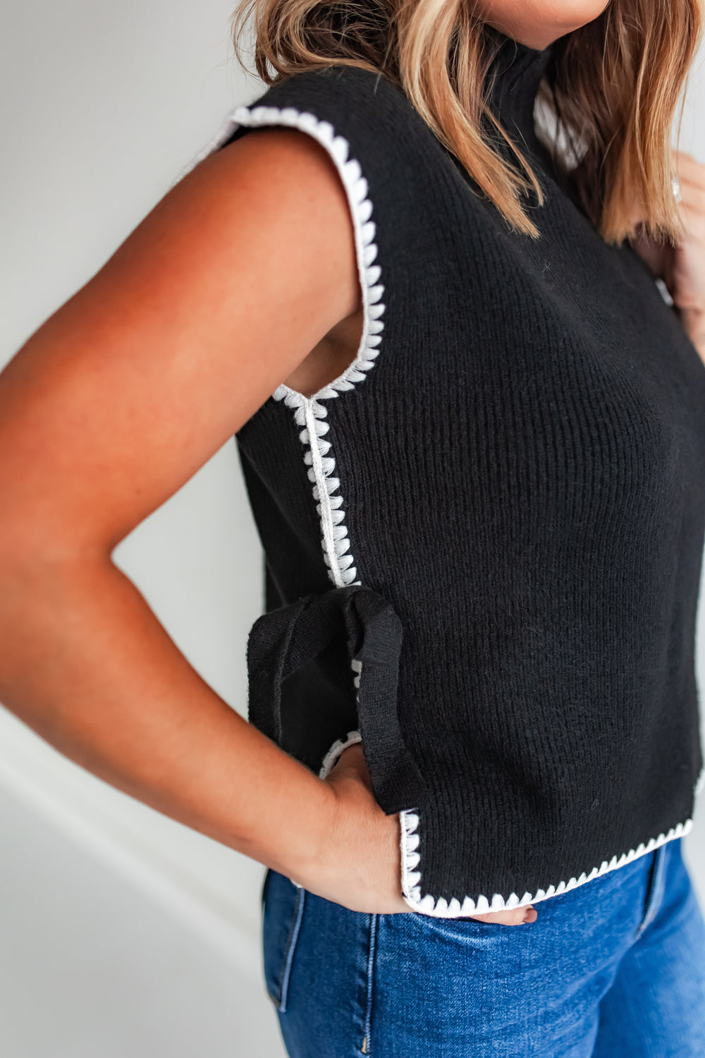 Woman wearing a black sleeveless sweater vest with white trim and blue jeans.