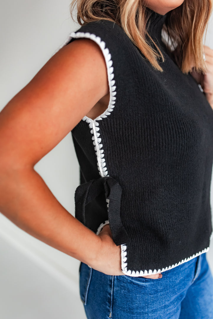 Woman wearing a black sleeveless sweater vest with white trim and blue jeans.