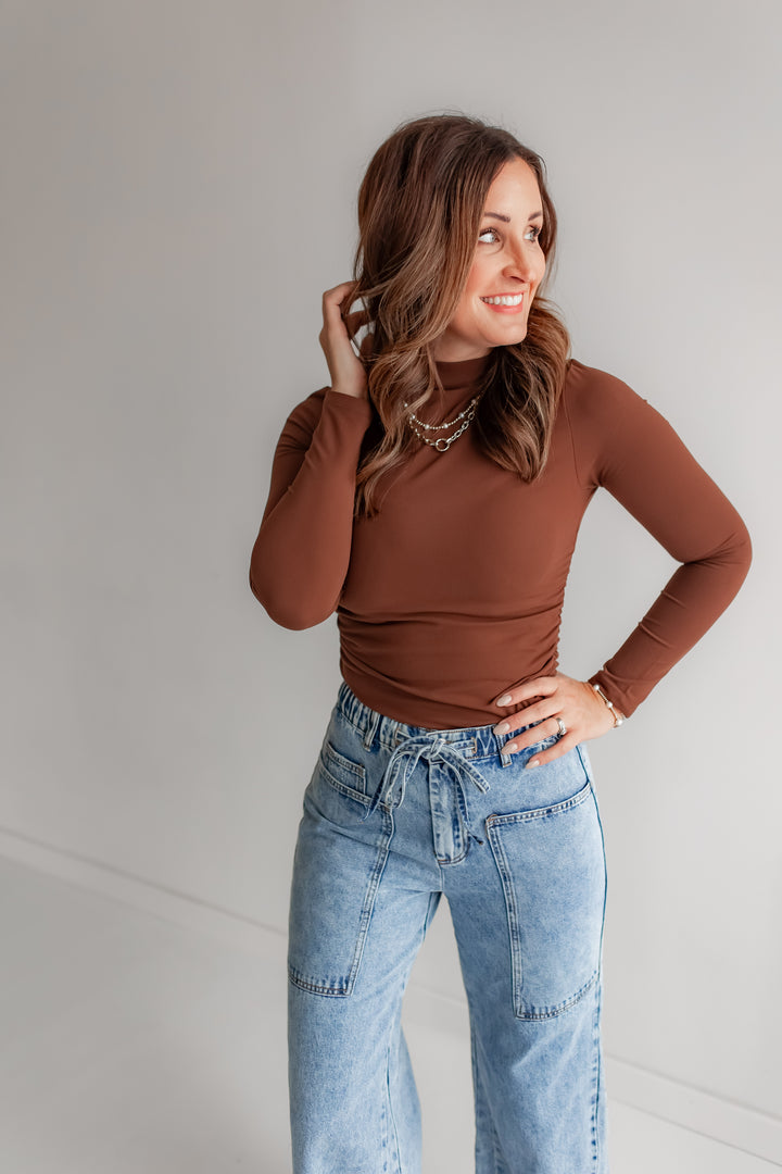 Woman wearing a brown long-sleeve top and cinching on the side with blue jeans 