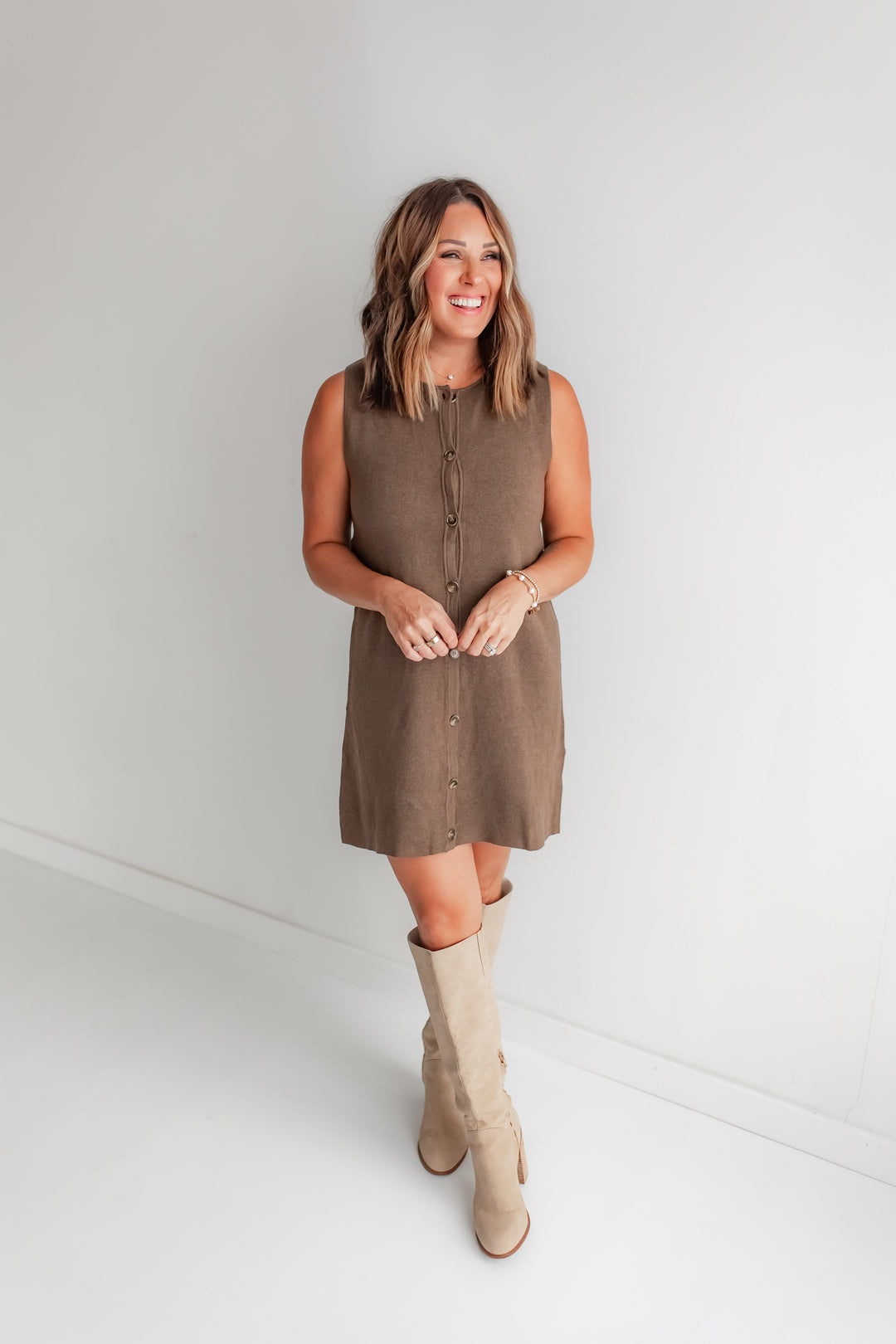 Woman wearing a brown sleeveless sweater dress and beige boots against a white background