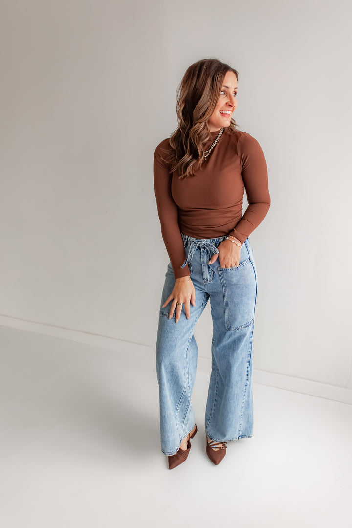 Woman wearing a brown top and light blue jeans with deep pockets and tie detail 