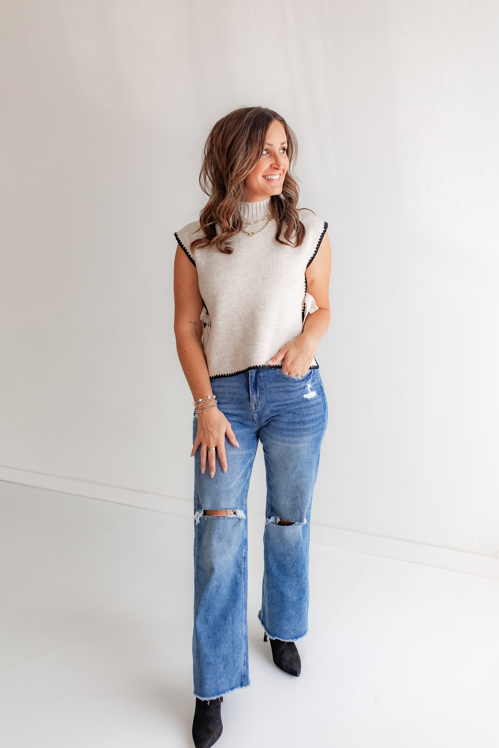 Model wearing ivory knit sleeveless top with black details and medium wash jeans and black booties