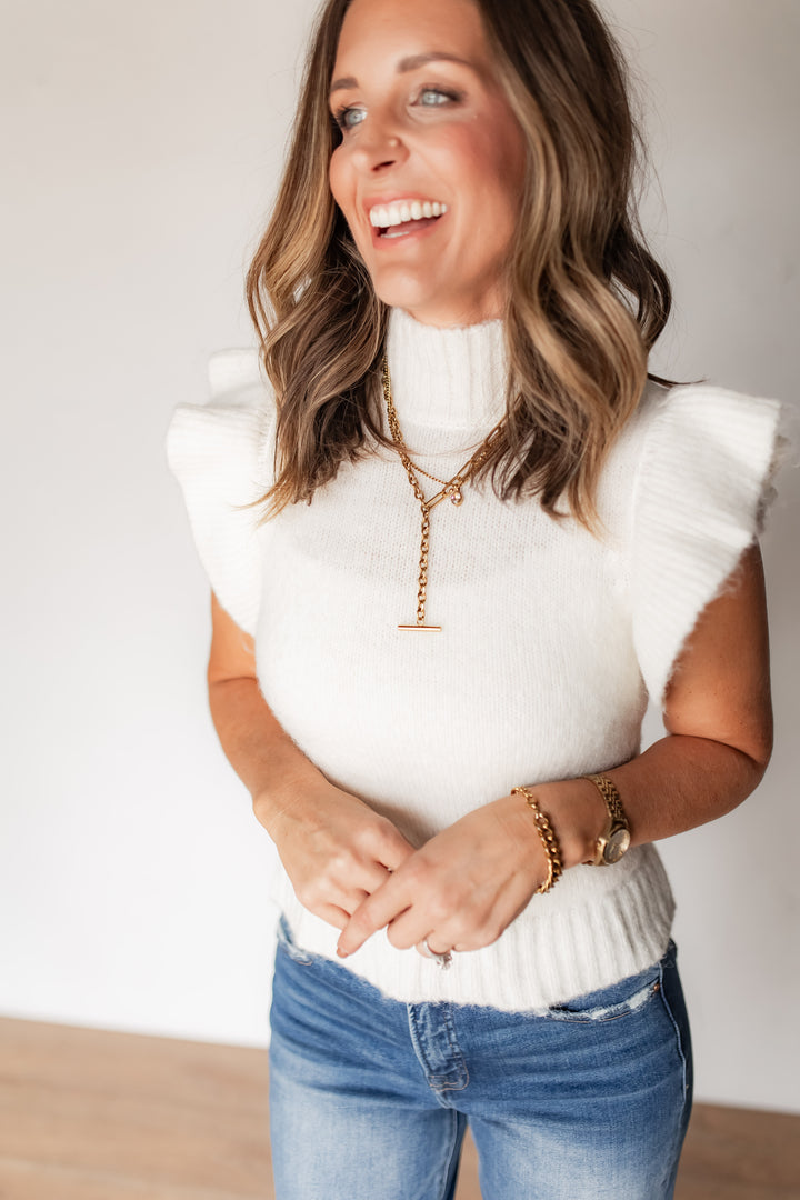 Woman wearing cream ruffle sleeve turtleneck sweater with gold layered necklaces and denim
