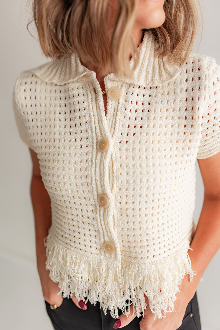 White crochet top with buttons and fringe on a plain background