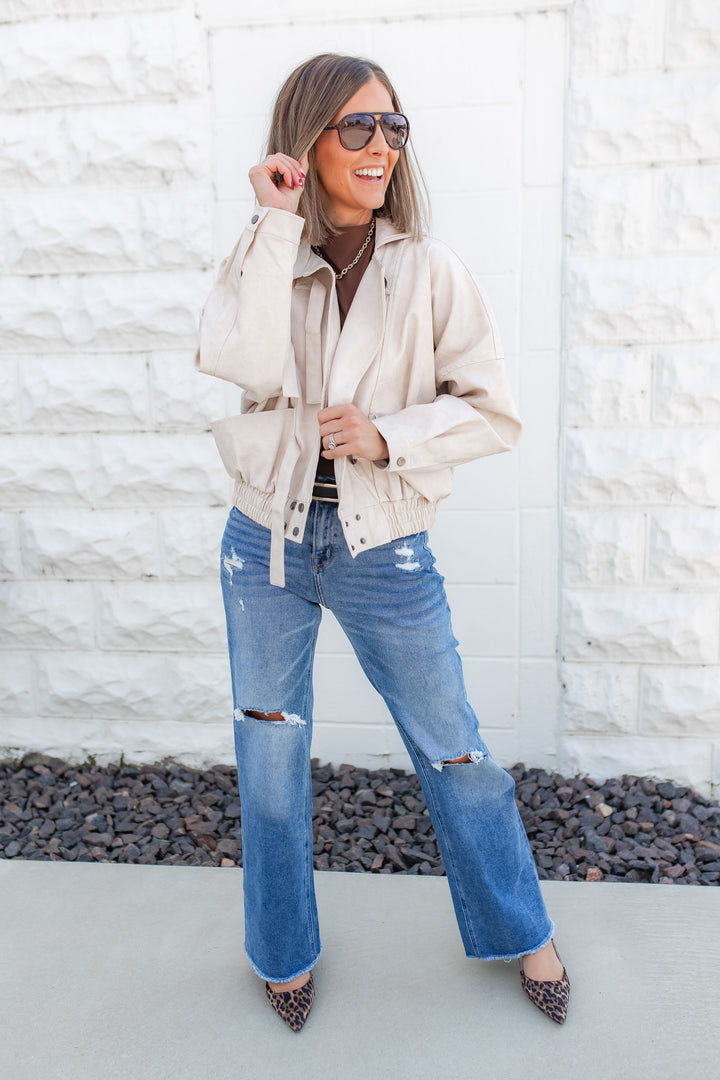 Woman wearing a beige jacket, blue jeans, and sunglasses against a white brick wall.