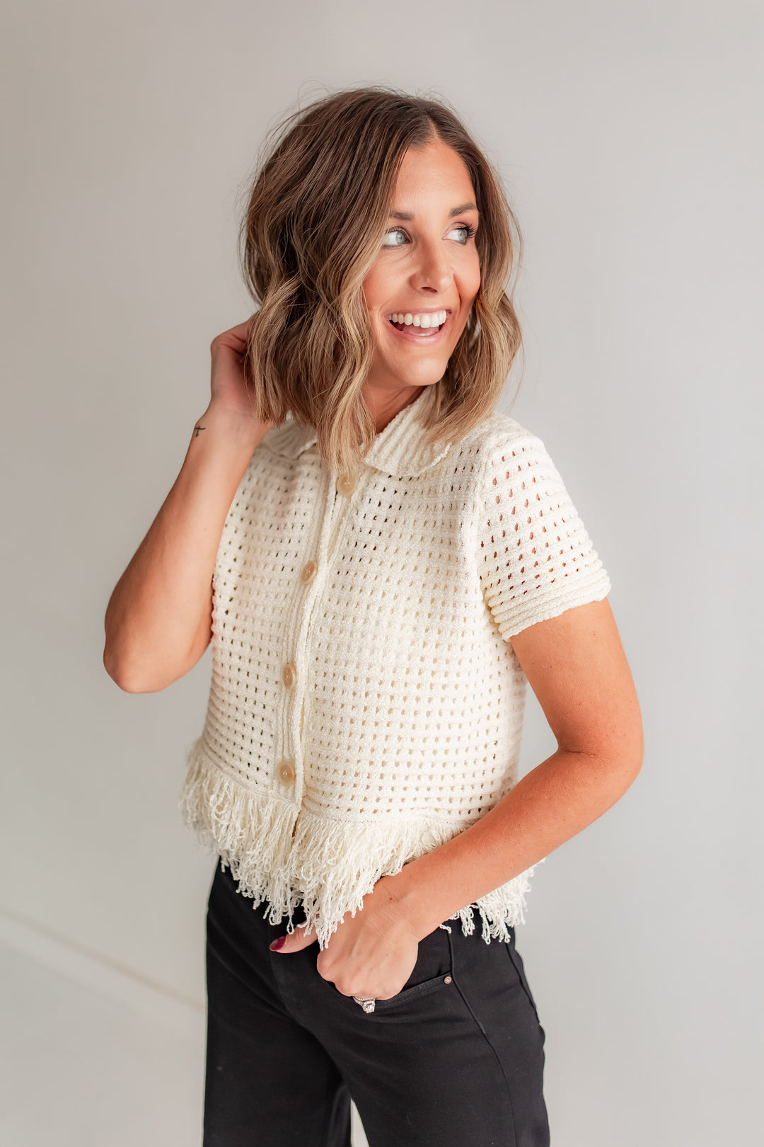 Woman wearing a white crochet top with fringe against a plain background