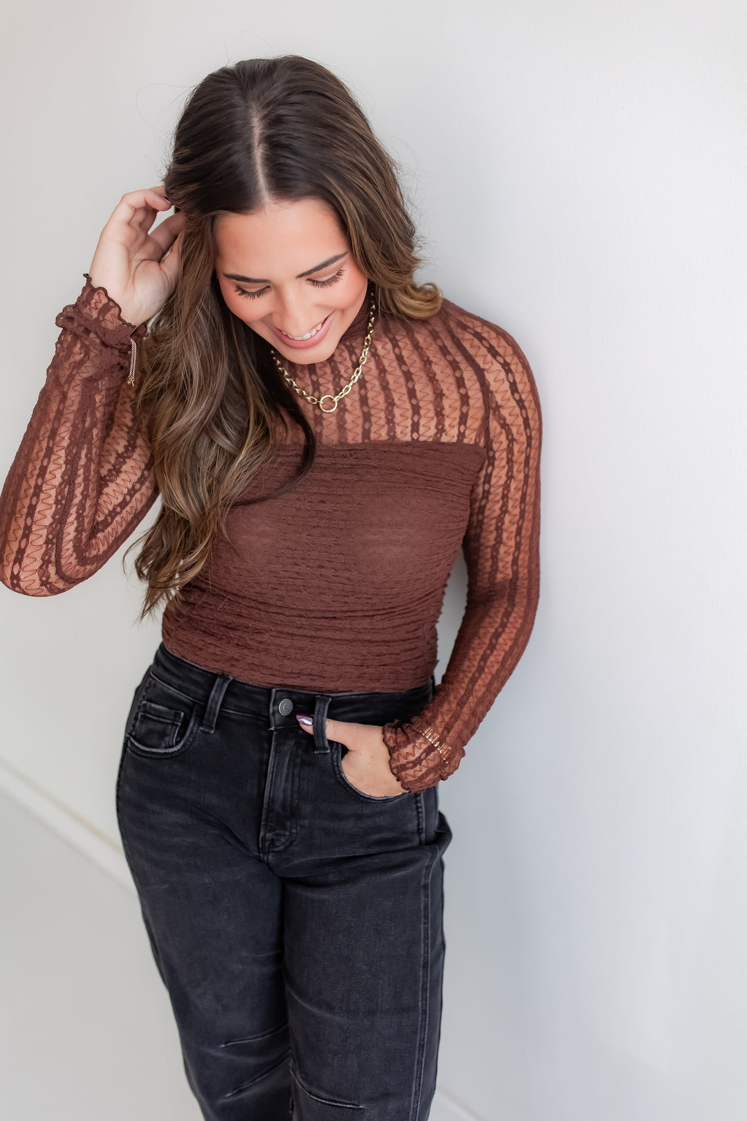Woman wearing a brown long-sleeve lace top and dark jeans against a white background