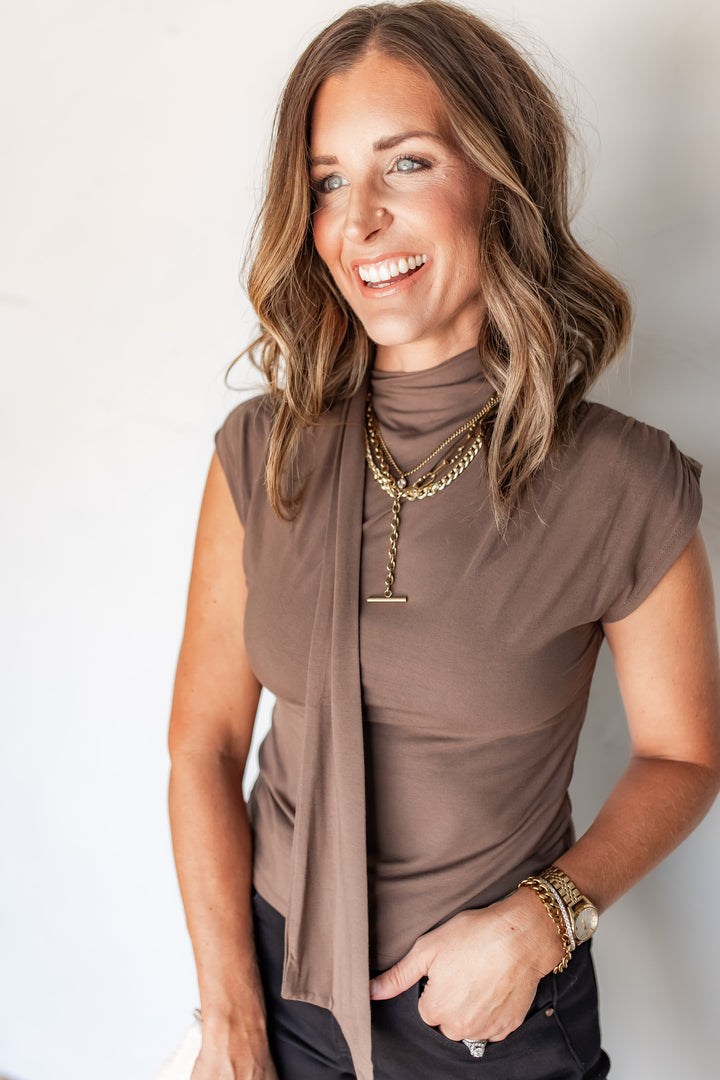 Woman wearing mocha knit top with draped scarf neckline and gold layered necklaces