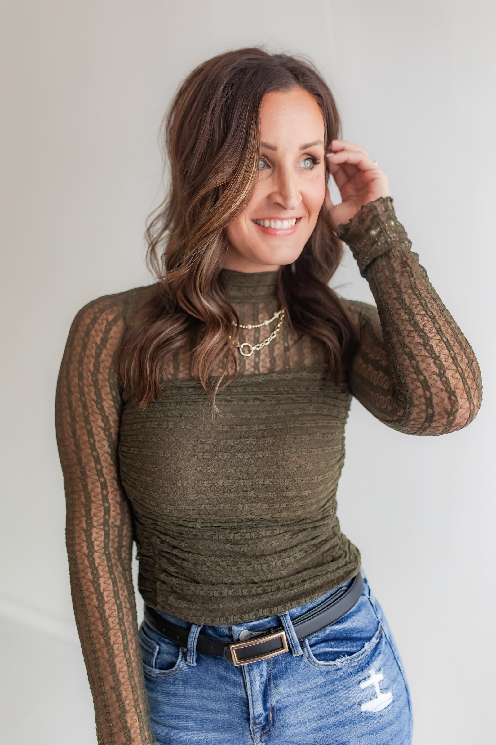Woman wearing a long sleeve lace bodysuit in a olive color and blue jeans against a plain background