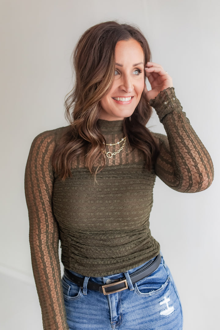 Woman wearing a long sleeve lace bodysuit in a olive color and blue jeans against a plain background