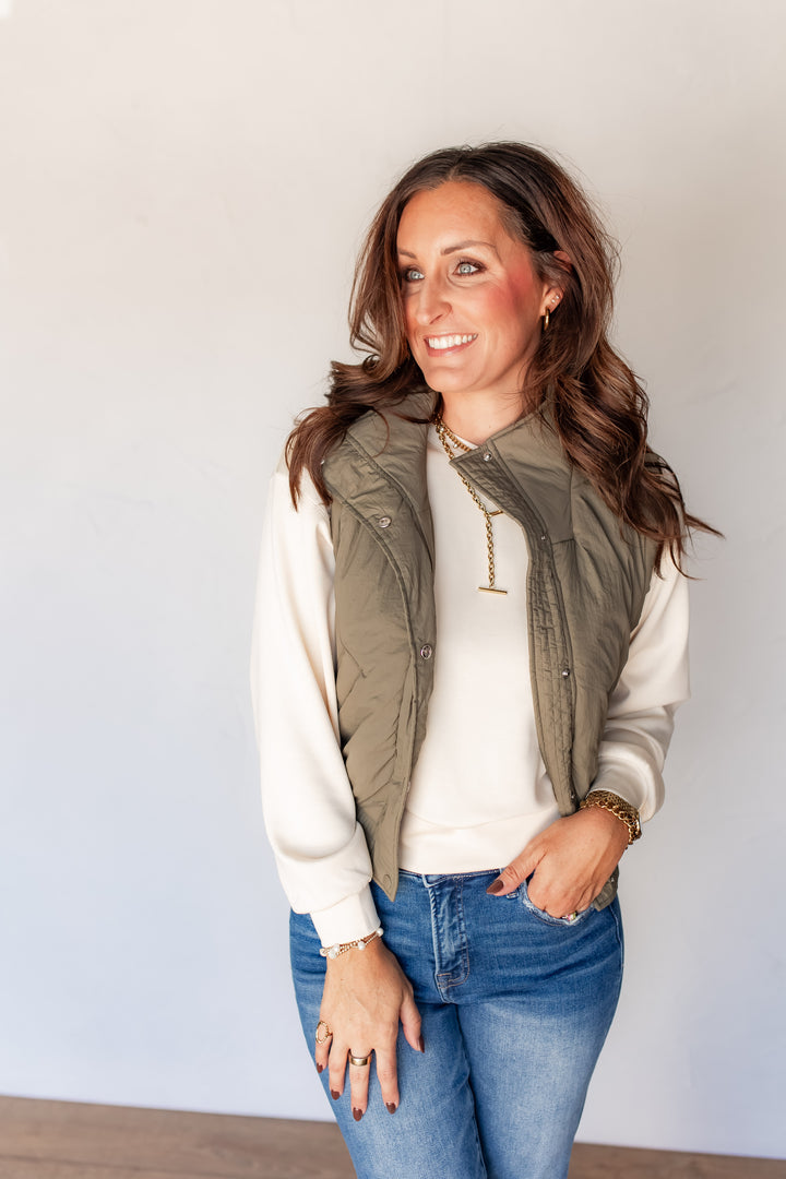 Woman wearing olive puffer vest with cream long sleeve pullover and medium wash jeans