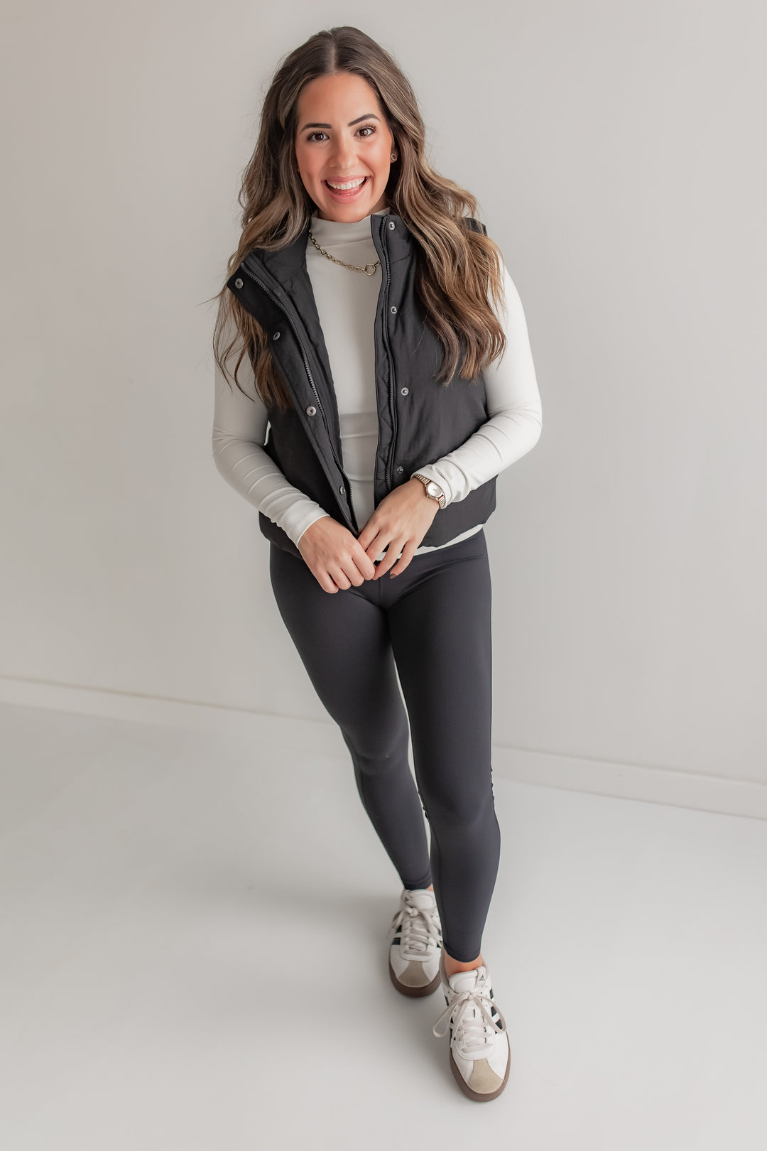 Woman wearing a black puffer vest, white sweater, black leggings, and white sneakers on a plain background