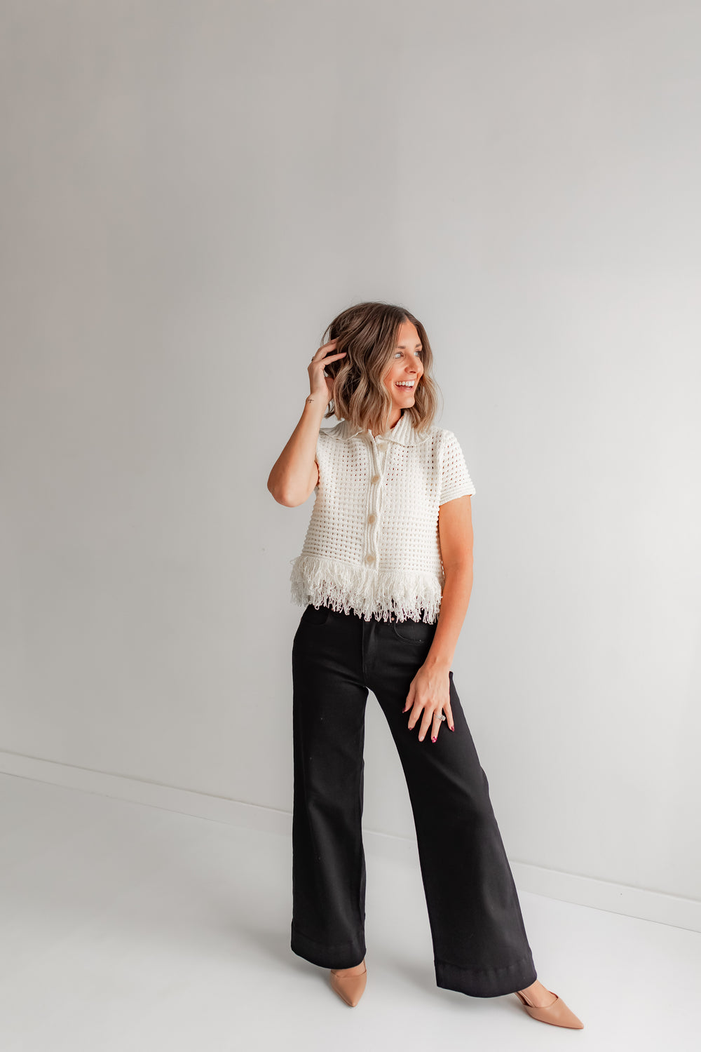 Woman wearing a short sleeve crochet sweater with fringe detail and black jeans. 