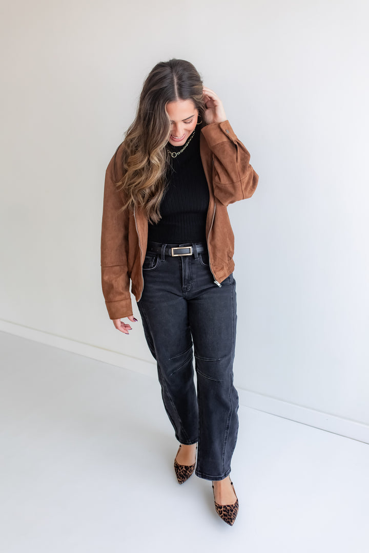 Woman wearing a brown jacket, black top, and dark jeans against a white background