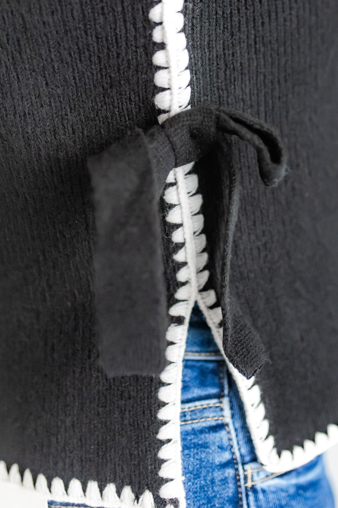 Close-up of a black sleeveless sweater with white stitching and ties, worn with blue jeans.