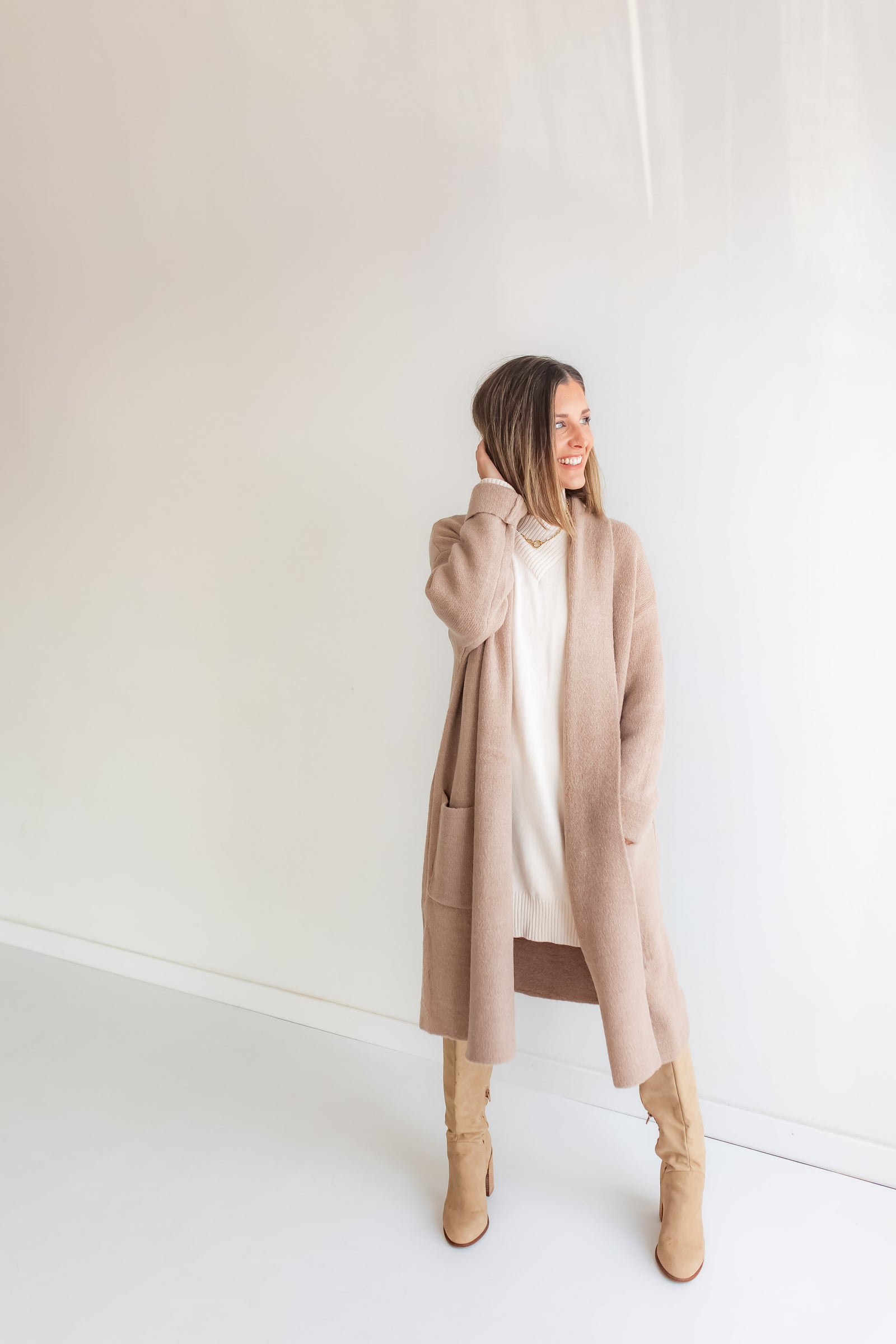 Woman wearing a long beige cardigan over a cream sweater dress against a white background