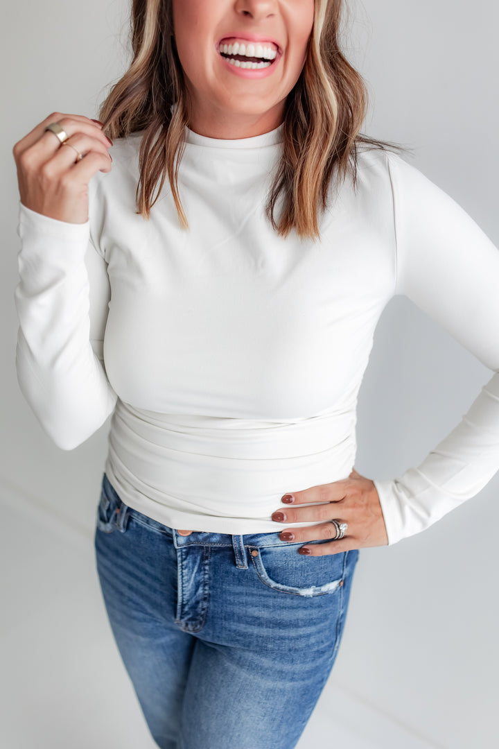 Woman wearing a white long-sleeve top and blue jeans against a plain background