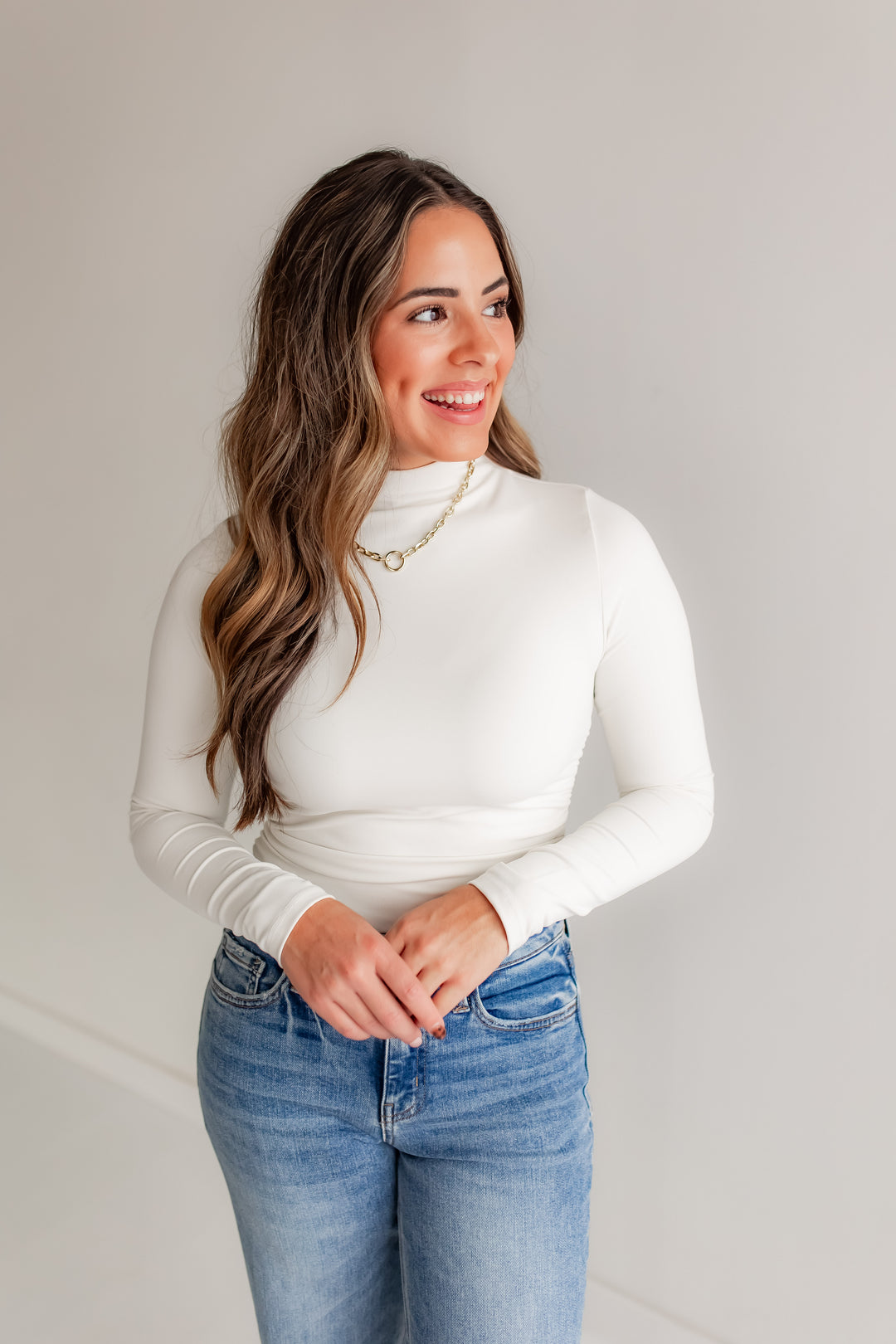 Woman wearing a white mockneck and blue jeans against a plain background