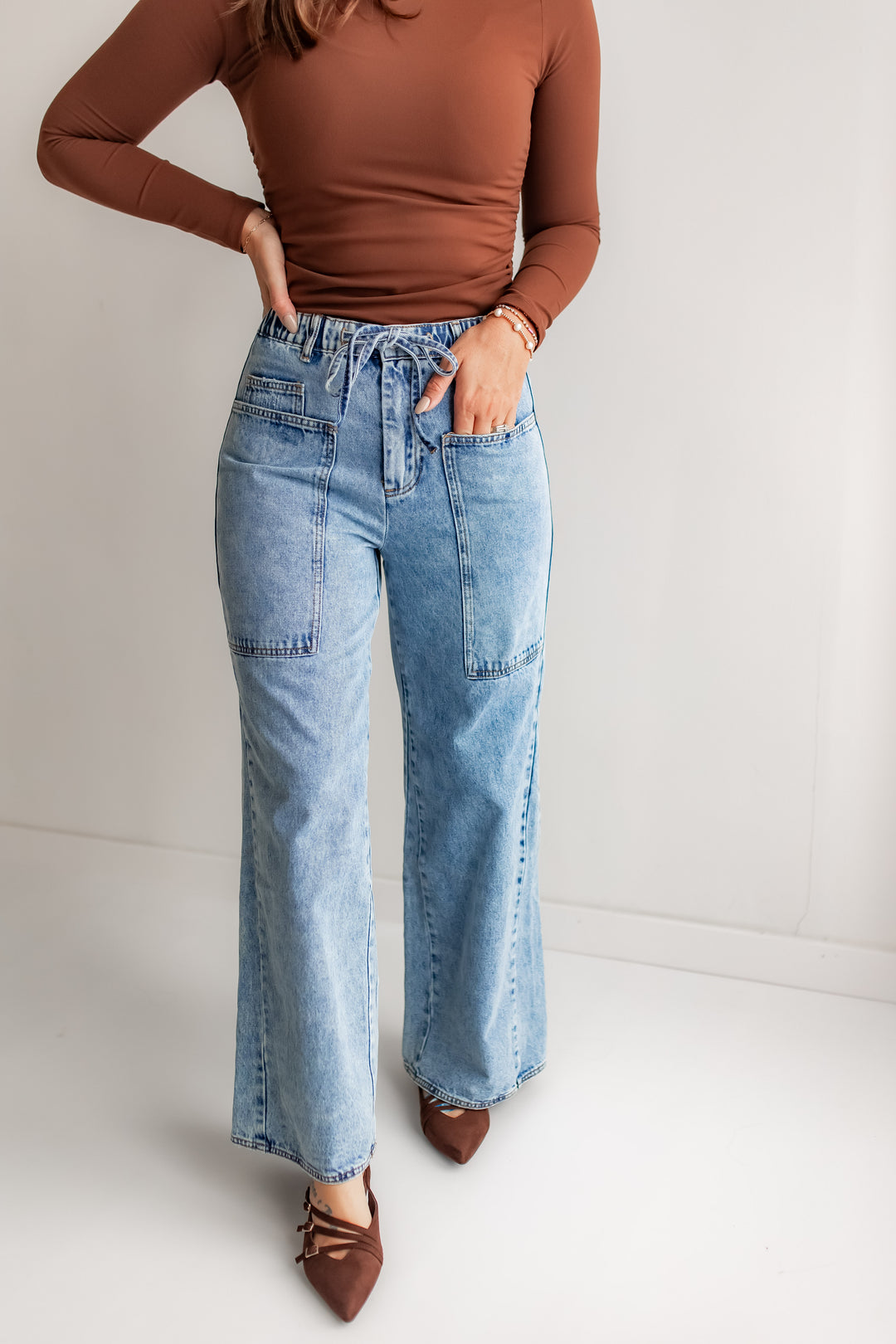 Woman wearing a brown top and blue wide leg jeans with a cinched waist and tie detail on a plain background